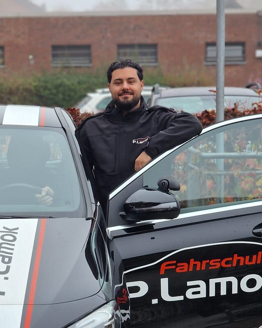 Mann lächelt neben schwarzem Auto mit Fahrschule-Logo, steht auf gepflasterter Fläche.